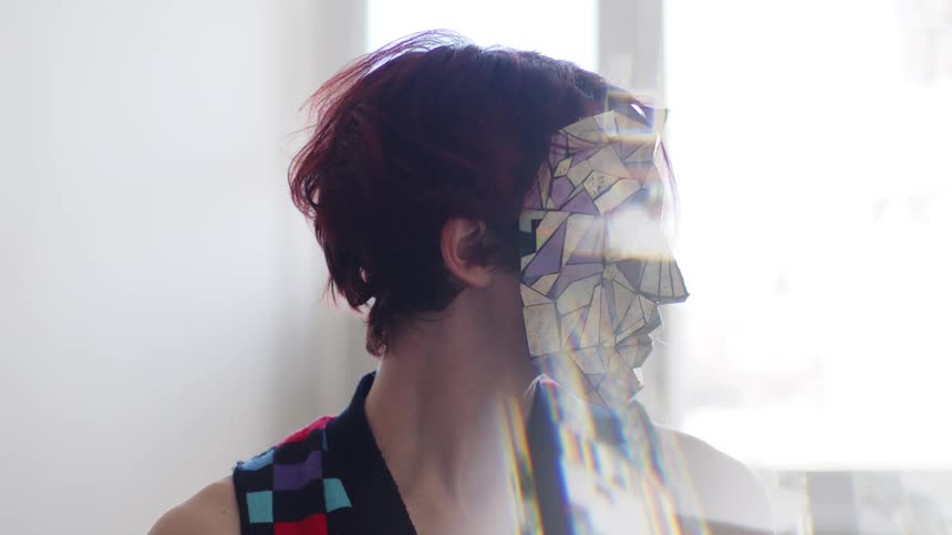 In a close up shot with a bright lens flare effect a woman with short red hair wears a mask made of reflective multi colored geometric shards as she slowly turns her head from side to side