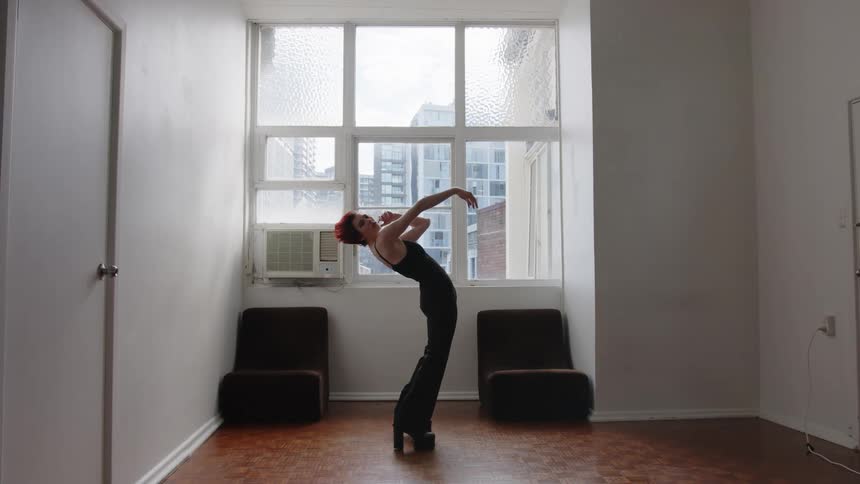 Woman dances with one arm raised and body arched in front of a window in an indoor space with buildings in the background