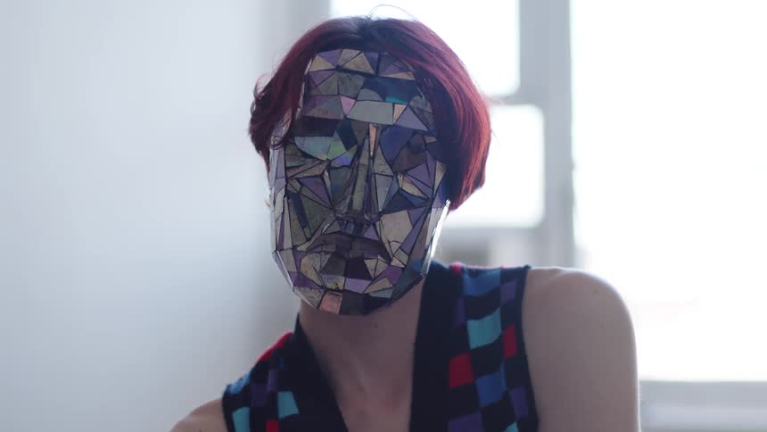 In a bright indoor space a person with short red hair wearing a checkered vest and a mask made of reflective multicolored geometric shards moves their head and hands touching their shoulders and face while looking toward and away from a nearby window