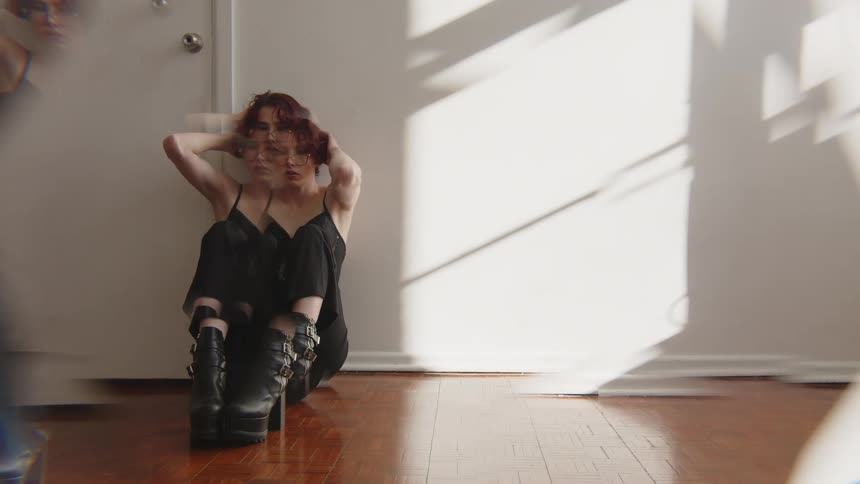 Woman sitting on the floor rests her back against a wall and places her hands on her head under natural light indoors