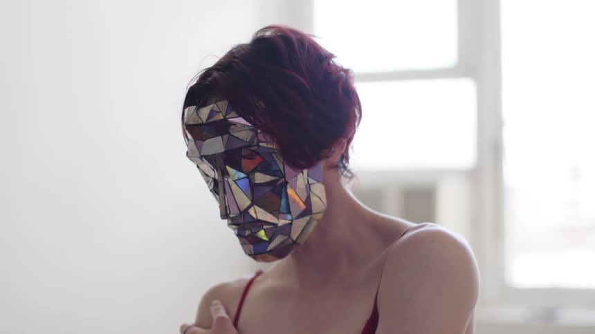 A person with short red hair wears a mask made of reflective geometric shards and a thin strapped top while moving their arms and hands gracefully against a bright blurred city background