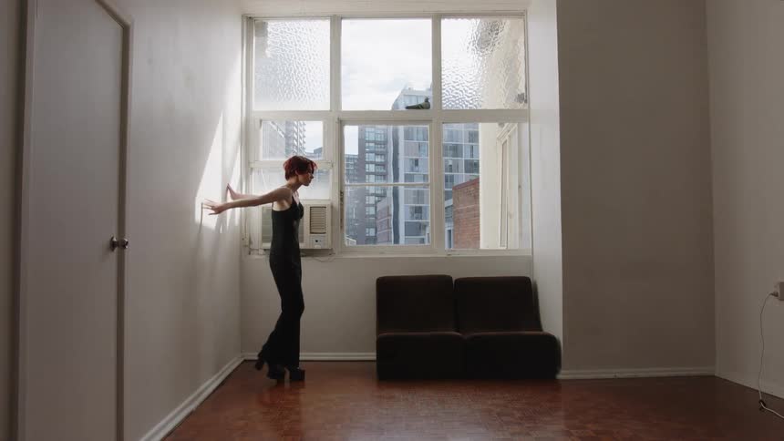 Woman standing in front of a window raises one arm over her head in an indoor space with buildings in the background