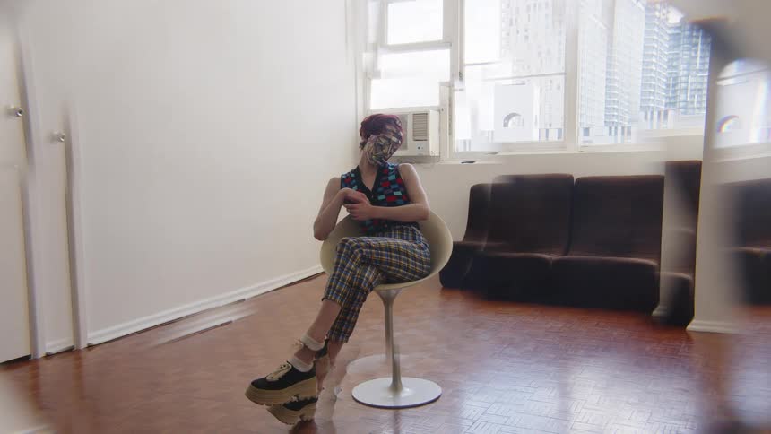 In a medium shot with a kaleidoscopic visual effect a woman wearing a red dress white boots and a reflective mosaic mask poses gracefully while reclining in a white chair within a sunlit room