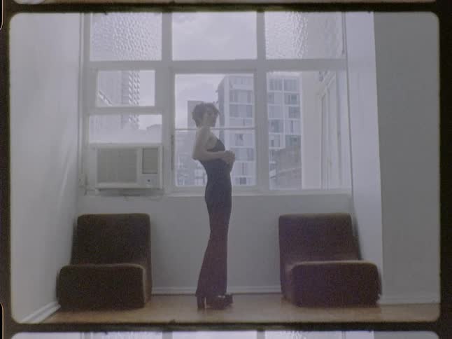 A woman with short red hair wearing a dark sleeveless shirt and platform boots poses in a sunlit room with white walls and a large window overlooking city buildings