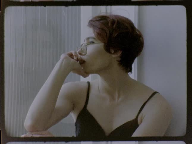 A woman with short red hair wearing glasses and a dark tank top sits by a window with a thoughtful expression in a close view captured with a vintage film aesthetic