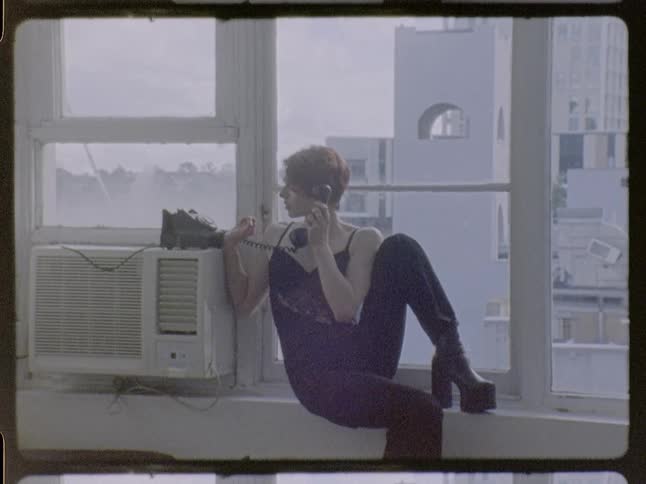 A woman with short red hair wearing a dark jumpsuit and platform boots sits on a windowsill and talks on a vintage telephone in a room overlooking city buildings
