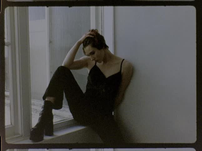 Woman sitting on a window ledge rests her hand on her forehead wearing black clothing indoors