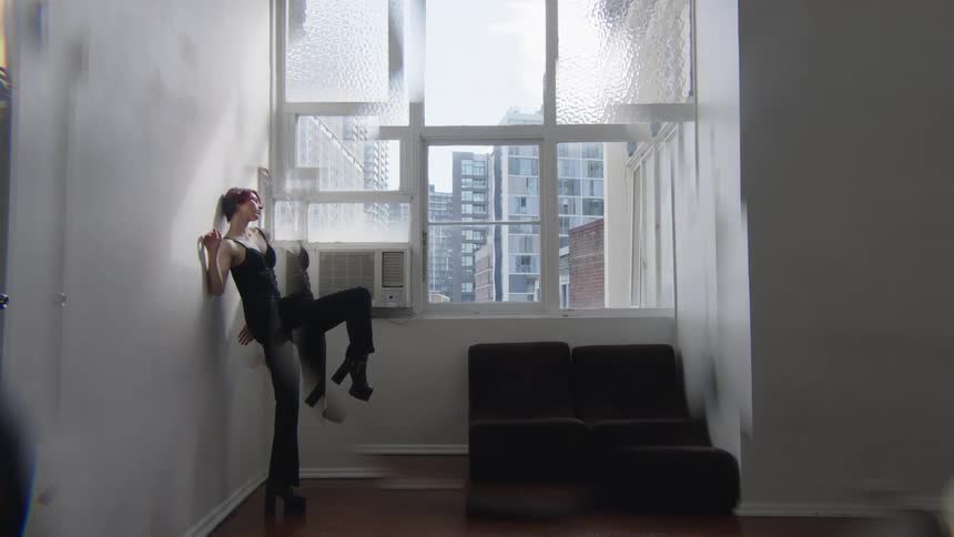 Woman leaning against a wall raises one leg in front of a window in an indoor space with buildings in the background