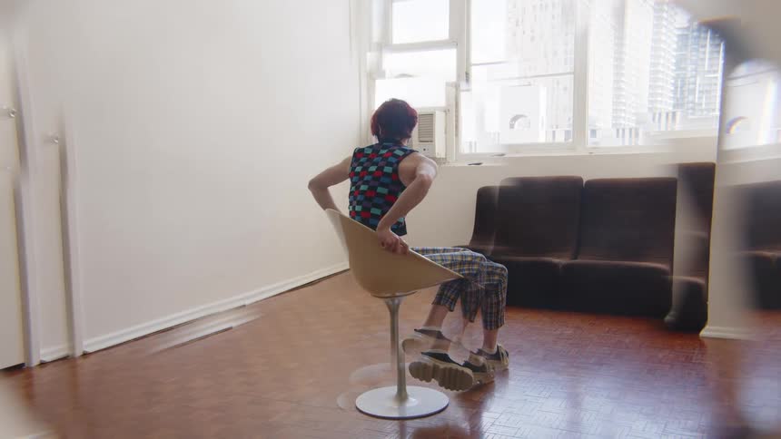 A woman with short red hair wearing a checkered vest and reflective mosaic mask sits in a white swivel chair and spins around in a sunlit room