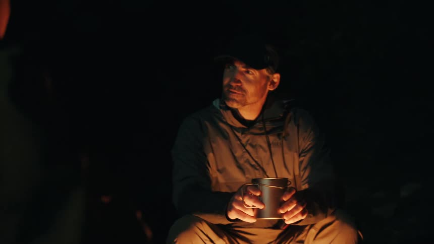 Man sitting in a dark environment holds a metal mug while making a hand gesture under warm light