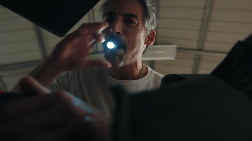 Man in an indoor environment holds a flashlight in his mouth while reaching toward an object out of frame