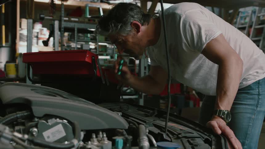 Mechanic wearing jeans and a white shirt leaning over the engine of a car in a garage indoors