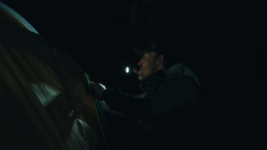 Man in a dark environment uses a headlamp while handling a tent at night