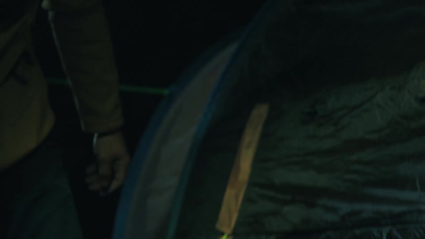 Young adult man wearing a yellow hacket and holding a flashlight on his mouth pitching a tent outdoors during night time