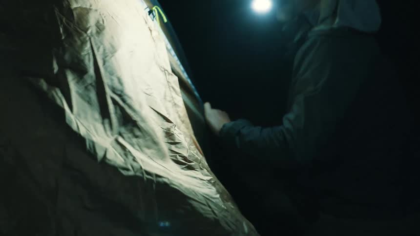 Bearded man wearing a baseball cap struggling to pitch a tent because of the wind outdoors during night time
