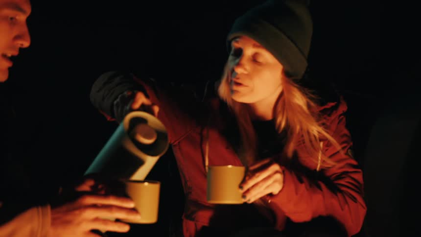 A woman wearing a beanie and jacket pours a hot beverage from a thermos for her male companion while sitting by a campfire at night