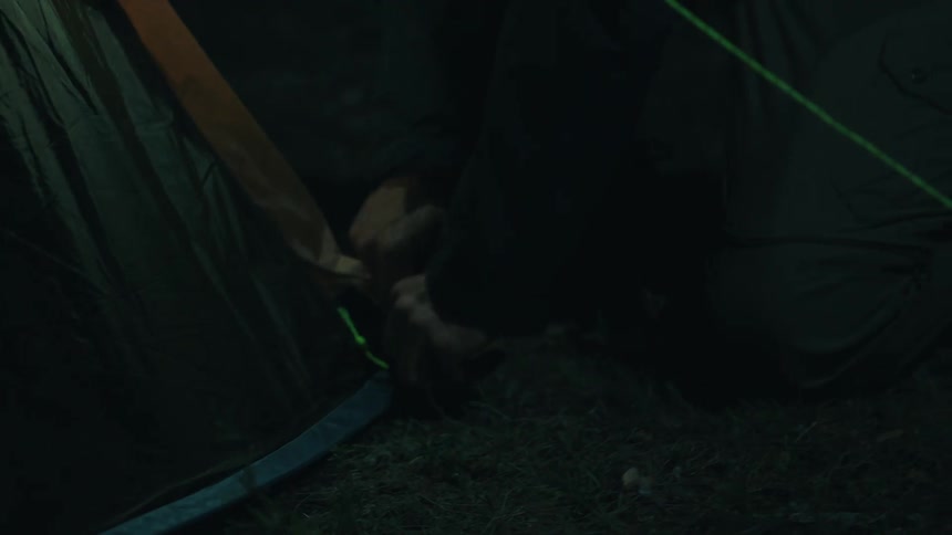 Adult man wearing a baseball cap and holding a flashlight on his mouth pitching a tent outdoors during night time