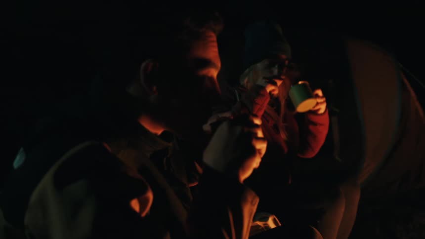 Friends drink from a mug while sitting in a campsite lit by firelight at night