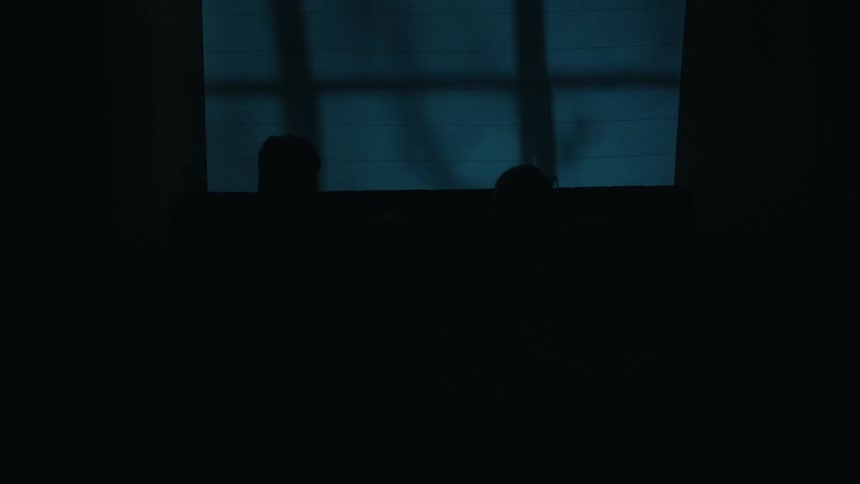 A man and woman sit in bed reading books and the light goes out transitioning the dimly lit room into complete darkness while shadows from a window are visible on the wall behind them