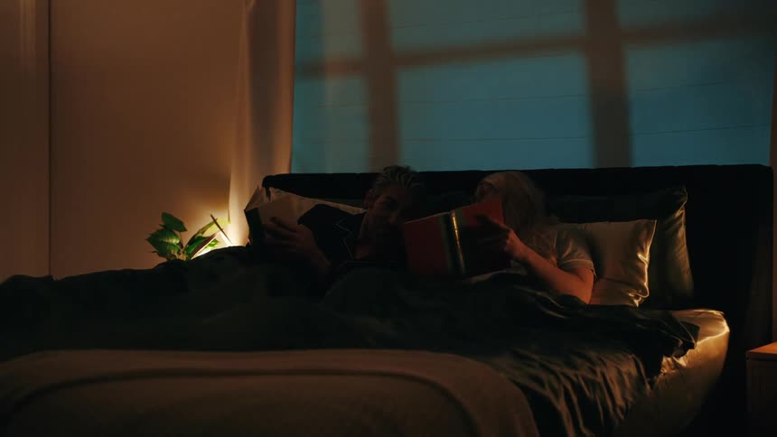 A man and woman read in bed under warm lamplight while looking forward with a quiet and relaxed mood