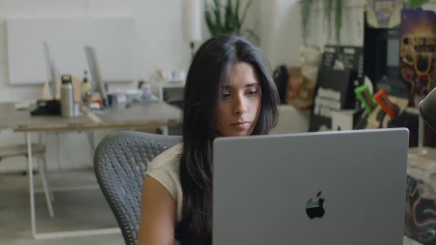 A young woman sits behind a desk in an office at midday typing on a computer keyboard as bright daylight fills the room