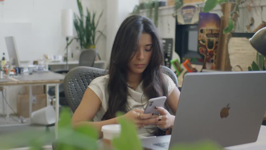 A young woman with long dark hair sits at a desk in a bright plantfilled office focusing on her smartphone while a laptop and a coffee cup rest in front of her