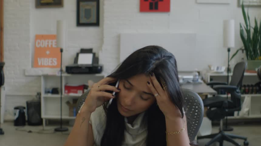 A stressed woman rubs her forehead while talking on a cell phone in an office