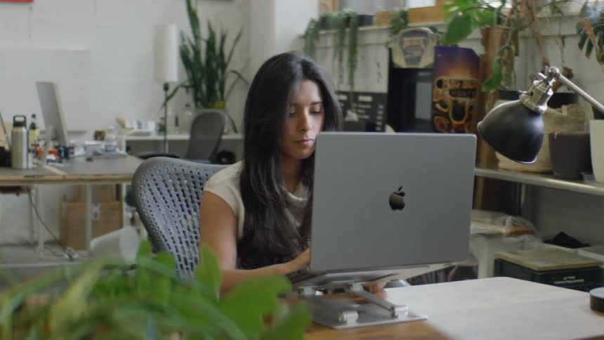 A woman with long dark hair sits at a wooden desk in a bright plantfilled office space focused on typing on a laptop that is propped up on a metal stand while she works in a quiet professional environment