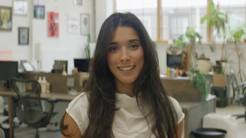 A woman with long dark hair wearing a white shirt looks into the camera and smiles in a professional office environment