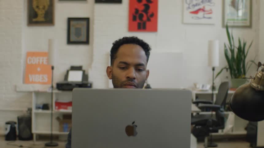 A man in a navy suit and white shirt uses a laptop in a bright office with posters looking forward with quiet concentration