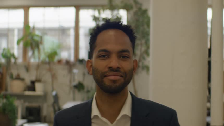 A man in a navy suit and white shirt stands in a bright office filled with plants looking forward with a calm and professional mood