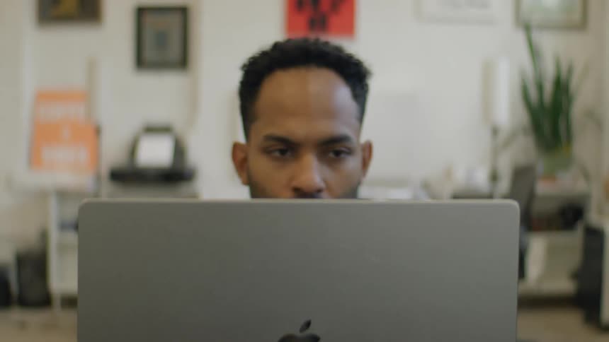 A man from his head up is shown looking at a laptop in deep thought in a home office room setting with posters and plants