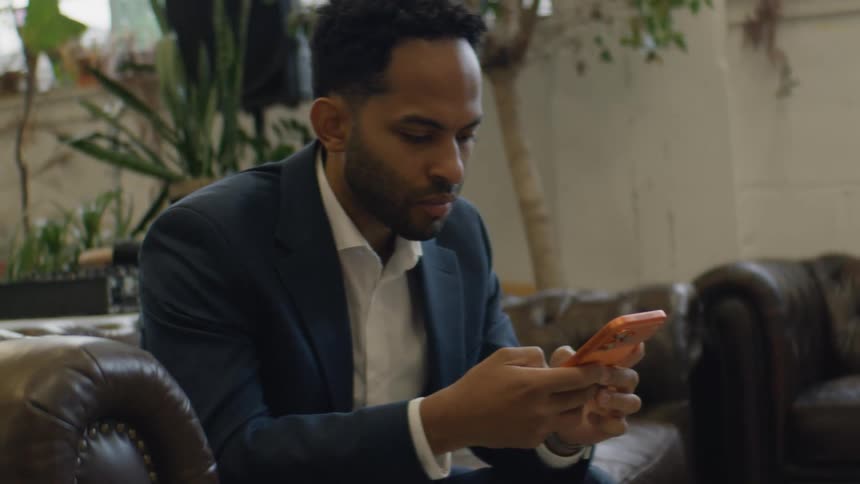 A young man sits on a couch at midday wearing a blue blazer and scrolling through his orange iphone 17