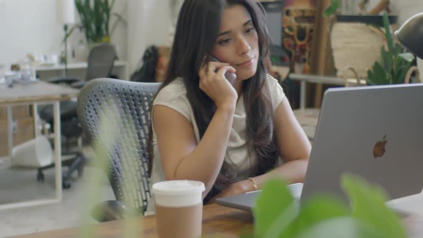 A woman sits at a desk in an office with a laptop open as she answers a smartphone holding it to her ear while looking at the screen and nodding during the conversation