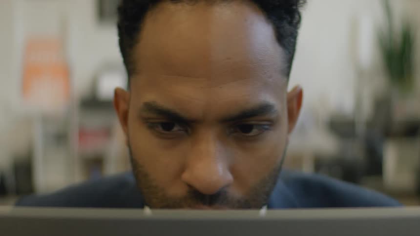Man in an indoor environment looks intently at a laptop screen in the foreground