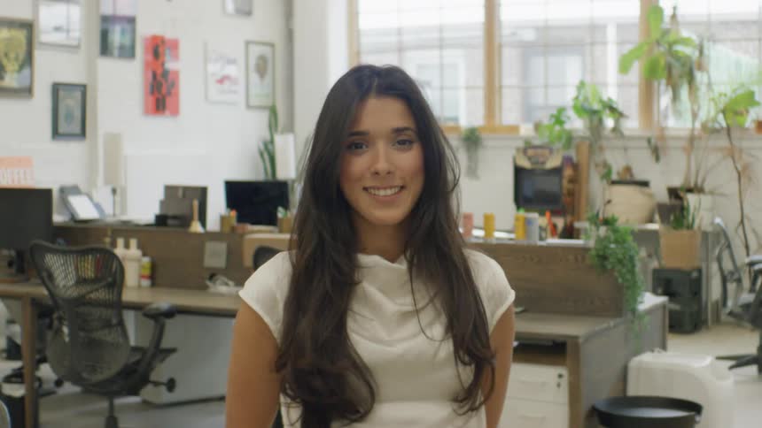 A woman with long dark hair wears a white sleeveless top in a bright office full of plants desks and pictures looking forward with a calm and professional mood