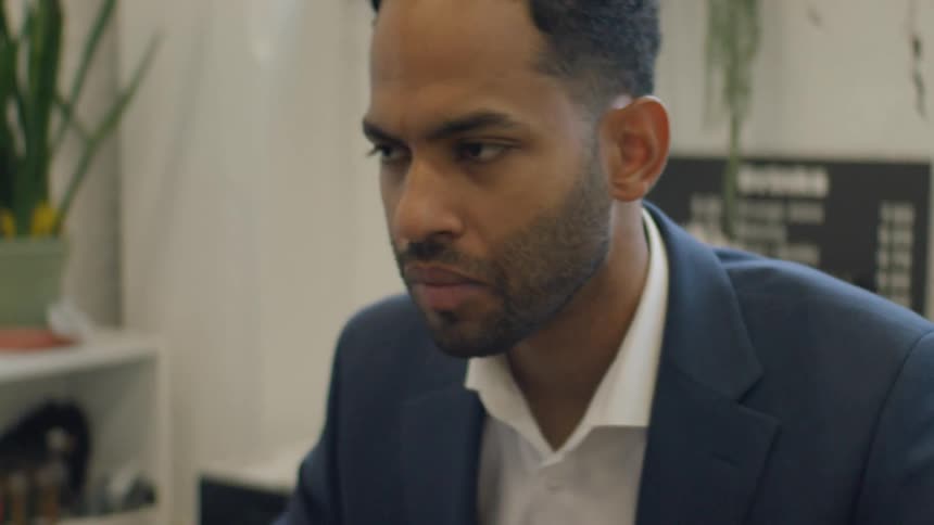 A worried man looks at a computer screen while sitting in a room with plants and a sign