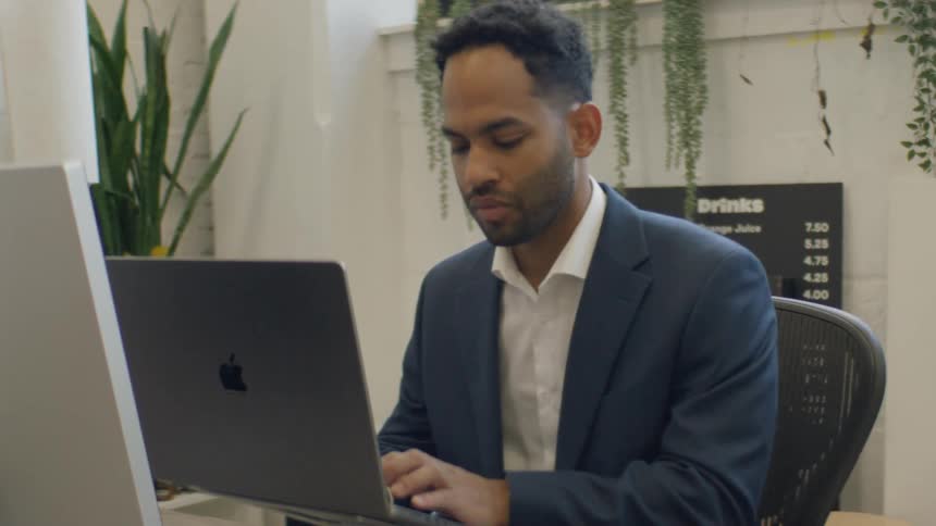 A man wearing a blue suit jacket and white shirt works at a laptop in a professional office environment