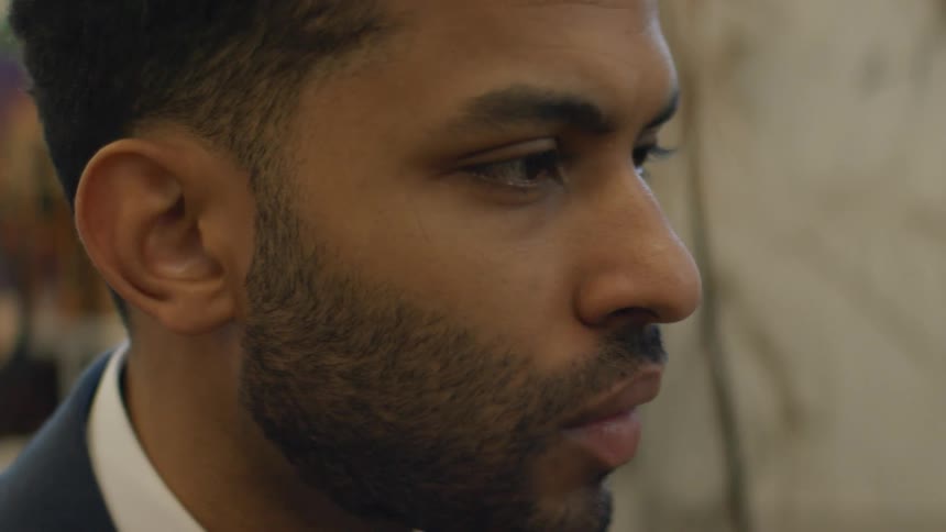 A man from the neck up in a navy suit and white shirt gazes thoughtfully to the side in a bright office environment looking forward with quiet concentration