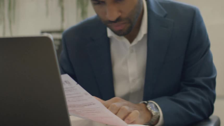 A man wearing a blue suit jacket and white shirt reviews a document while sitting at a desk with a laptop in a professional office