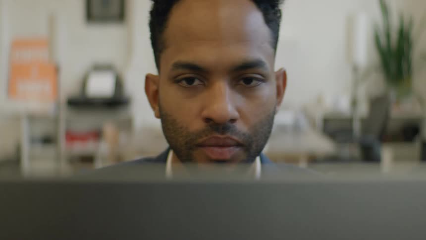 A man in a navy suit and white shirt leans forward over a laptop with his hand to his chin looking forward with quiet concentration
