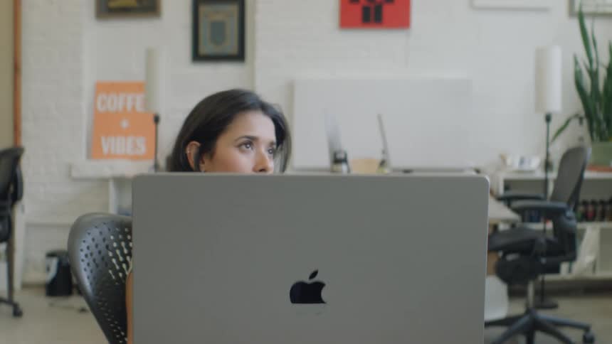 A woman with long dark hair uses a laptop in a bright office looking upward with quiet concentration