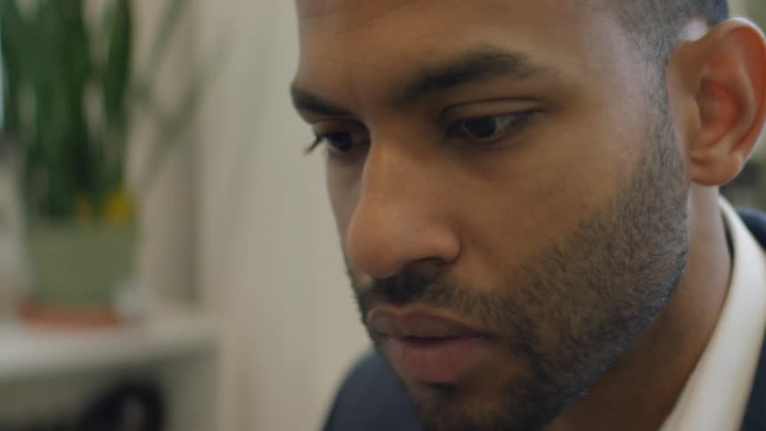 Young adult black man wearing a shirt and blazer sitting down with a nervous facial expression indoors during day time