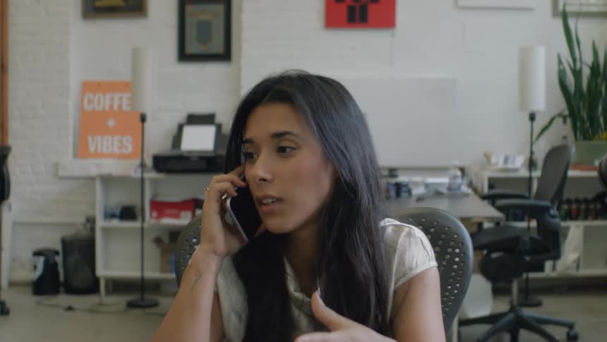 A woman with long dark hair wearing a white shirt speaks on a mobile phone while sitting in a professional office environment