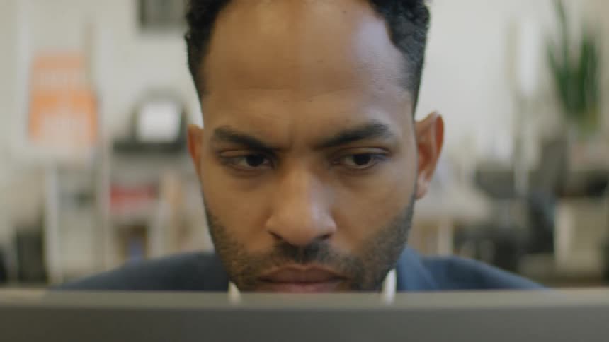 A man with a beard and dark hair wearing a blue blazer stares intensely at a computer screen while resting his hand on his chin in a brightly lit office environment displaying a focused and pensive expression