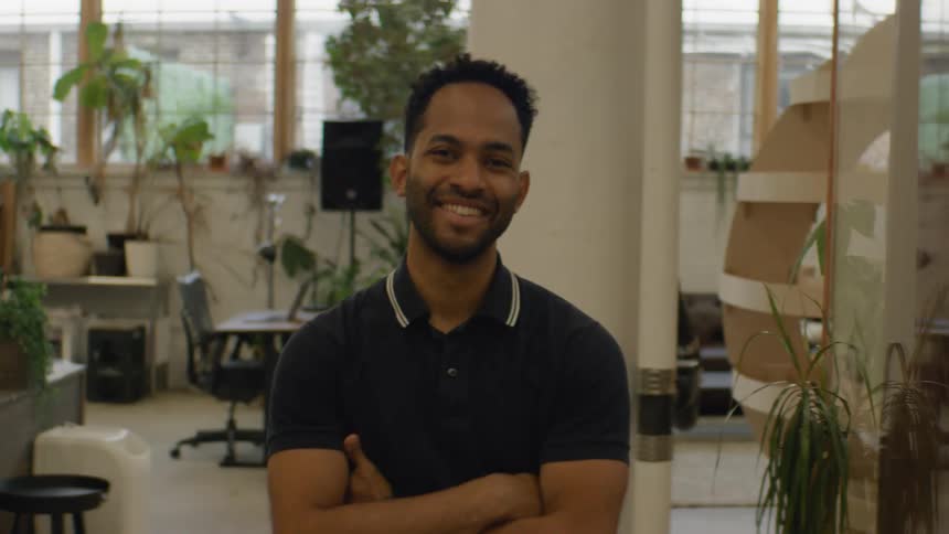 A man with a short beard and dark hair stands in an openoffice environment smiling at the camera before crossing his arms over his chest in a confident posture while various office plants and furniture blur into the background