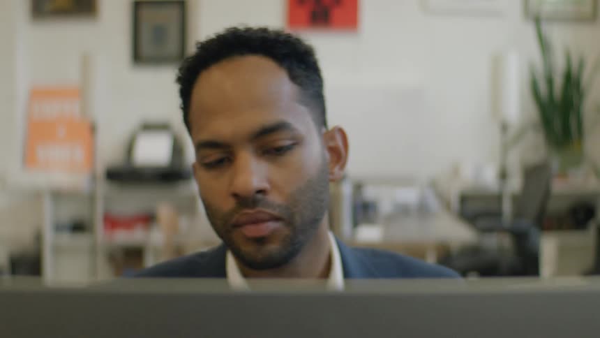A man wearing a blue suit jacket and white shirt works at a laptop in a professional office environment