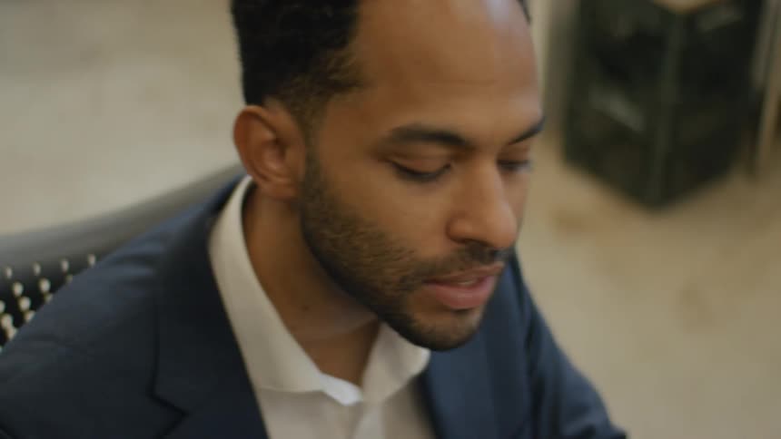 A frustrated man with a beard wearing a blue suit jacket and a white shirt speaks in a professional office setting
