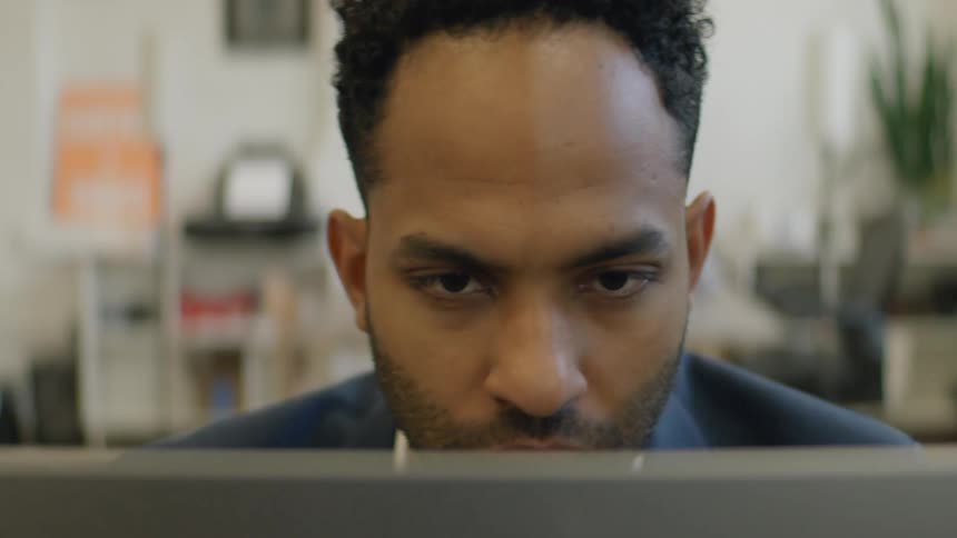 Young adult black man wearing a blazer sitting down in a room and reading on a laptop computer with a contemplative facial expression indoors during day time