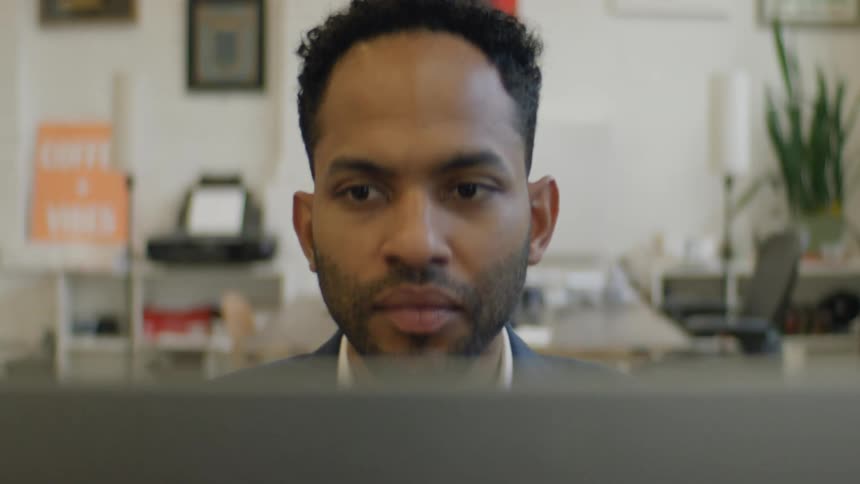 A man sits in an office setting intently focused on the screen of his laptopas the camera slowly zooms in his expression remains serious and concentrated on his work